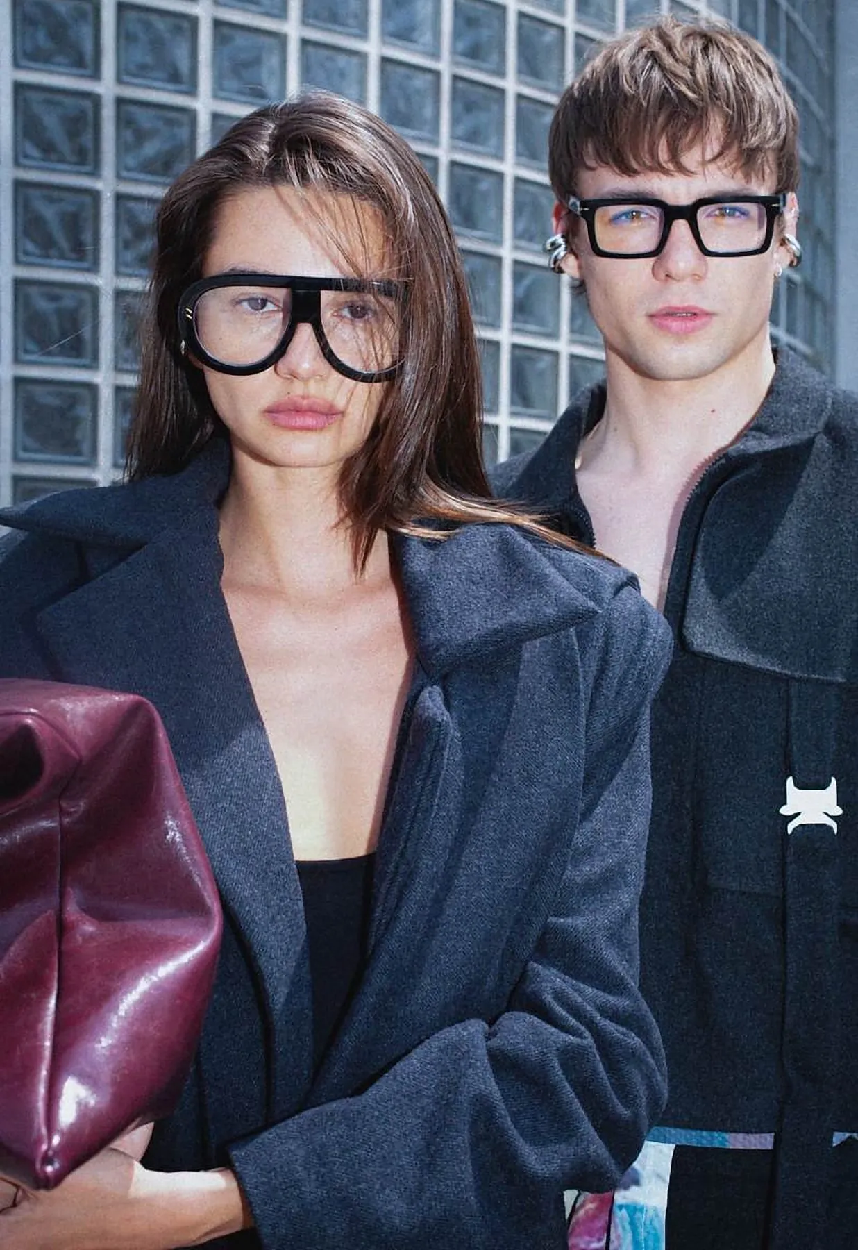 Models in an oversized grey wool coat and black jacket in front of a glass-brick wall