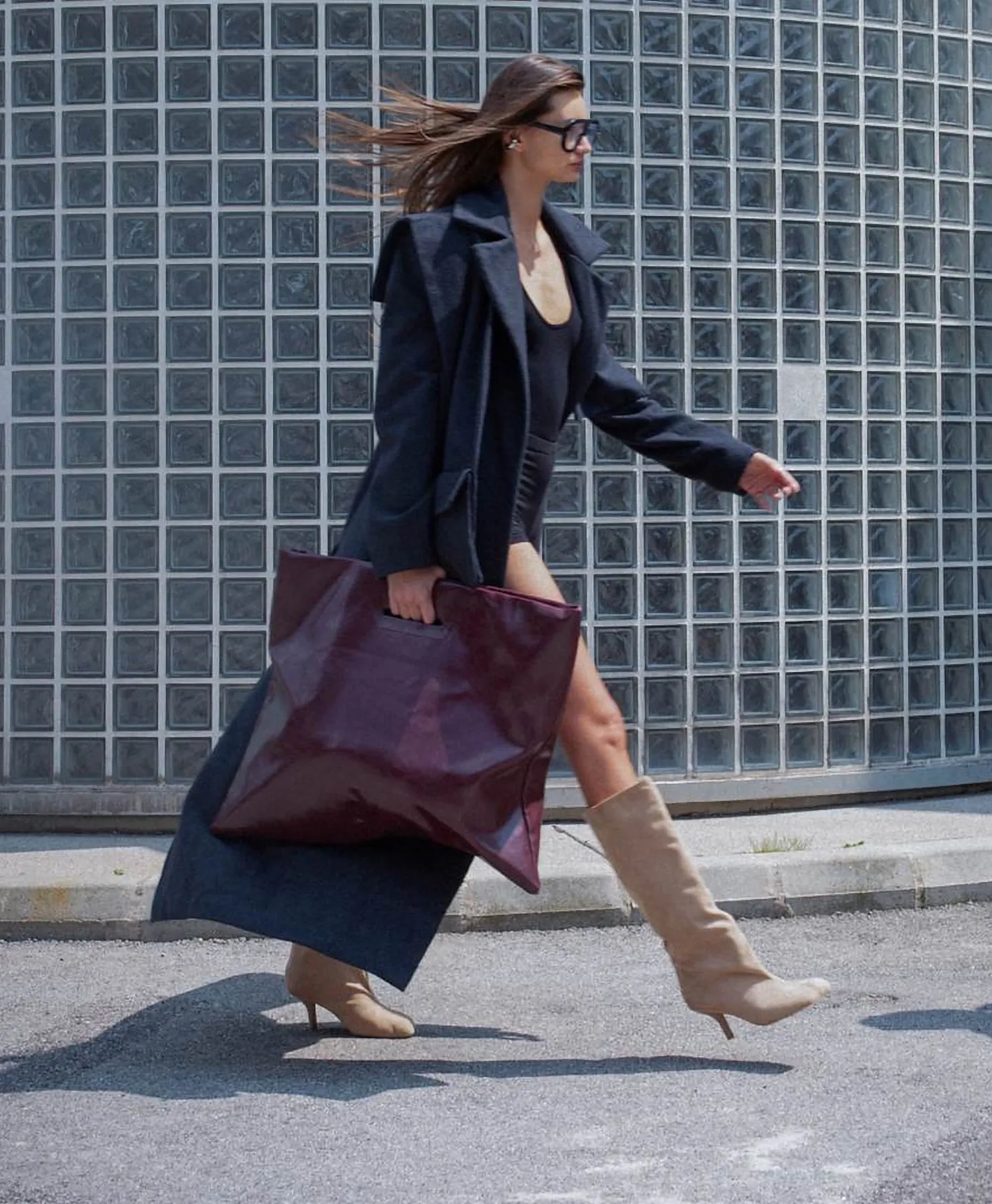 Model in a navy coat walking past a glass-brick wall with a burgundy bag