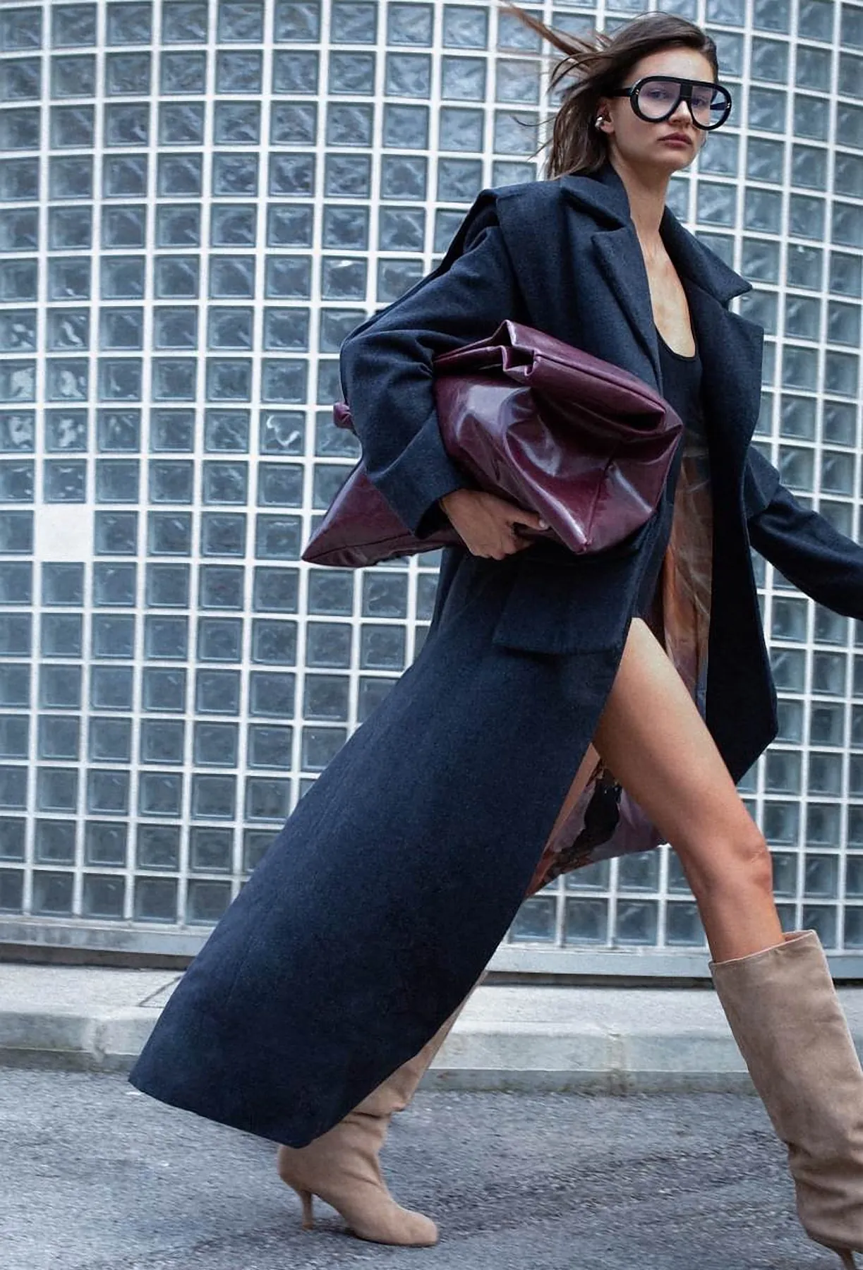 Model walking in a long navy coat carrying a burgundy leather bag