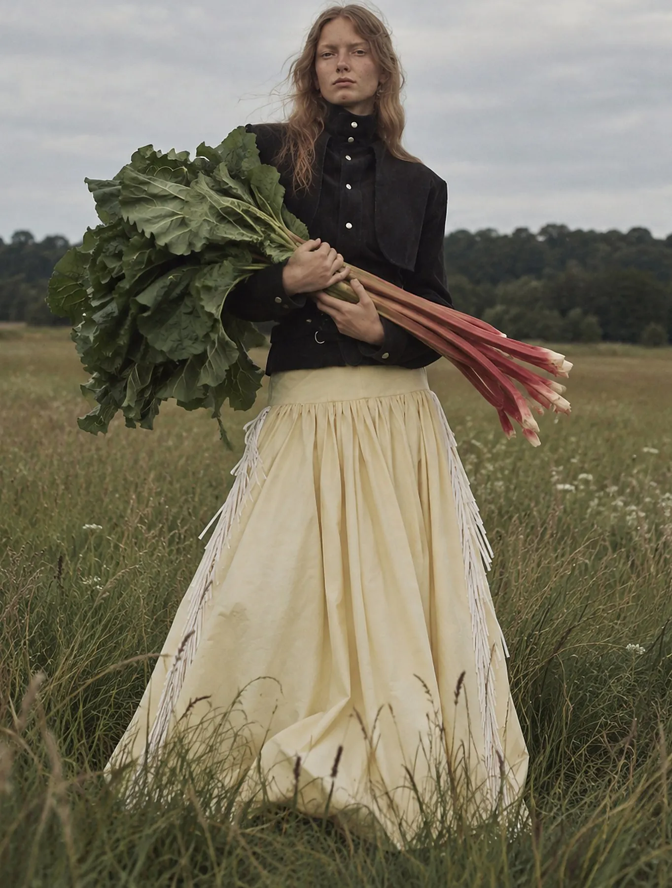 Model in black suede jacket and cream fringed skirt holding rhubarb in a field